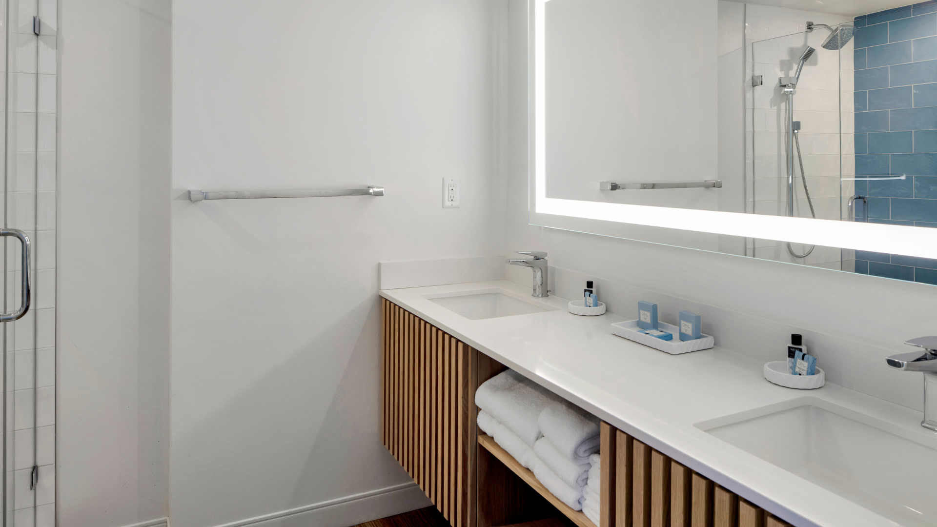 Modern bathroom with a large, illuminated mirror above a double sink vanity. The vanity features wooden slats and holds folded towels, toiletries, and soap dispensers. A glass shower with blue tiles is visible in the background.