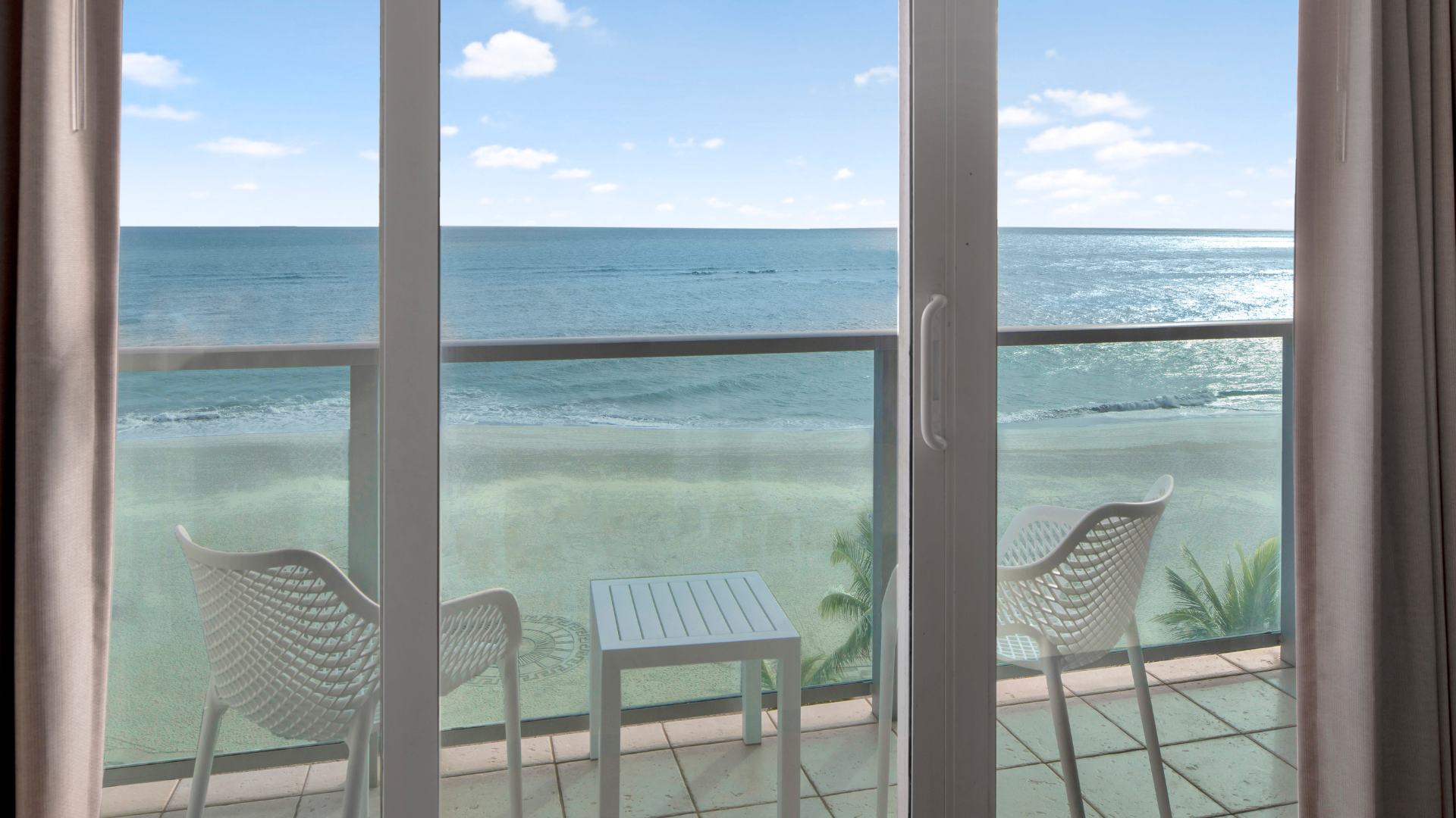 View from a balcony overlooking a sandy beach and ocean. Two white chairs and a small table are placed facing the water, framed by glass doors. The sky is clear with a few clouds.