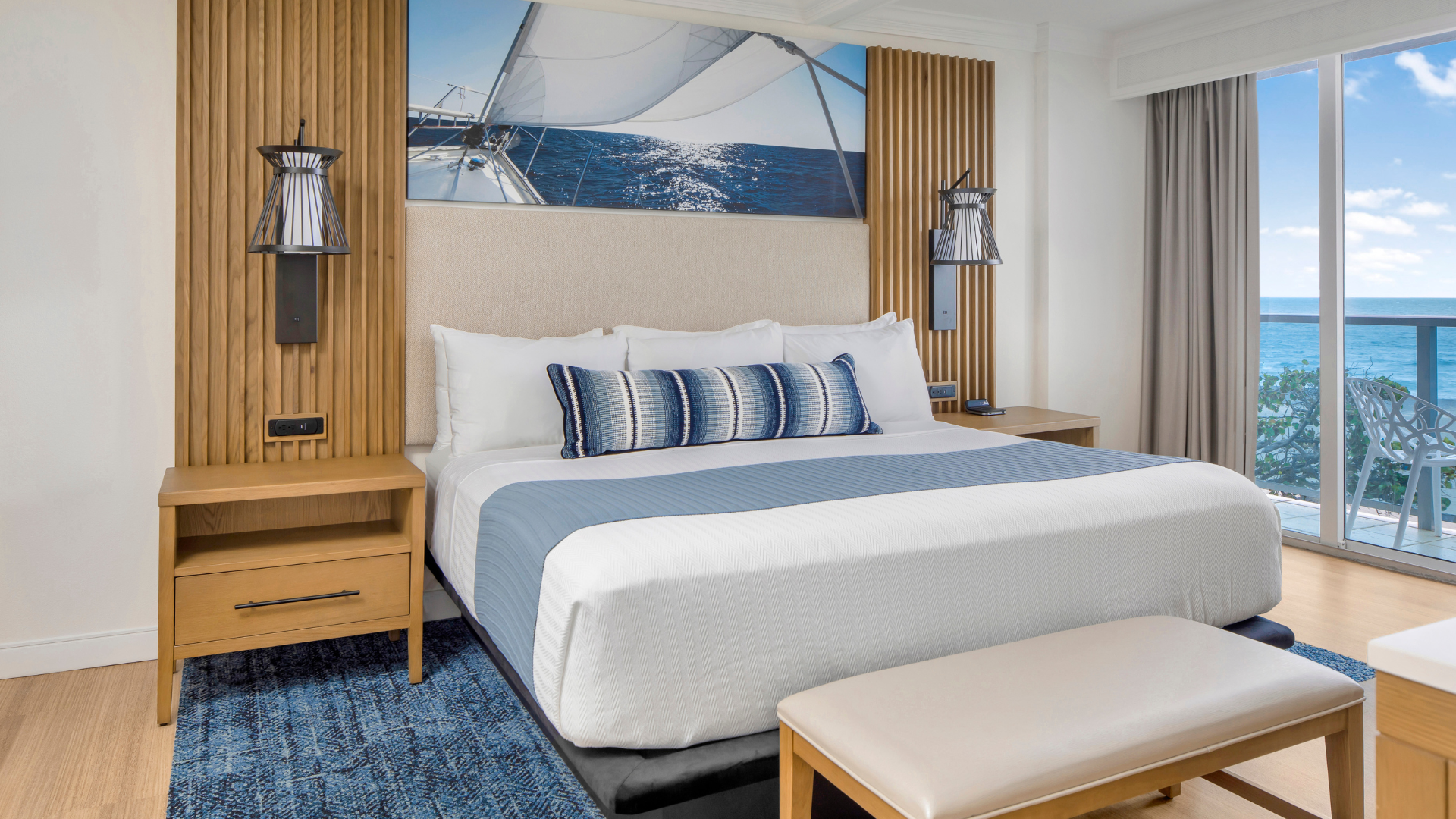 A bright hotel room features a neatly made bed with blue and white bedding, flanked by wooden nightstands and modern lamps. Theres a nautical-themed picture above the bed, and a glass door opens to a balcony with an ocean view.