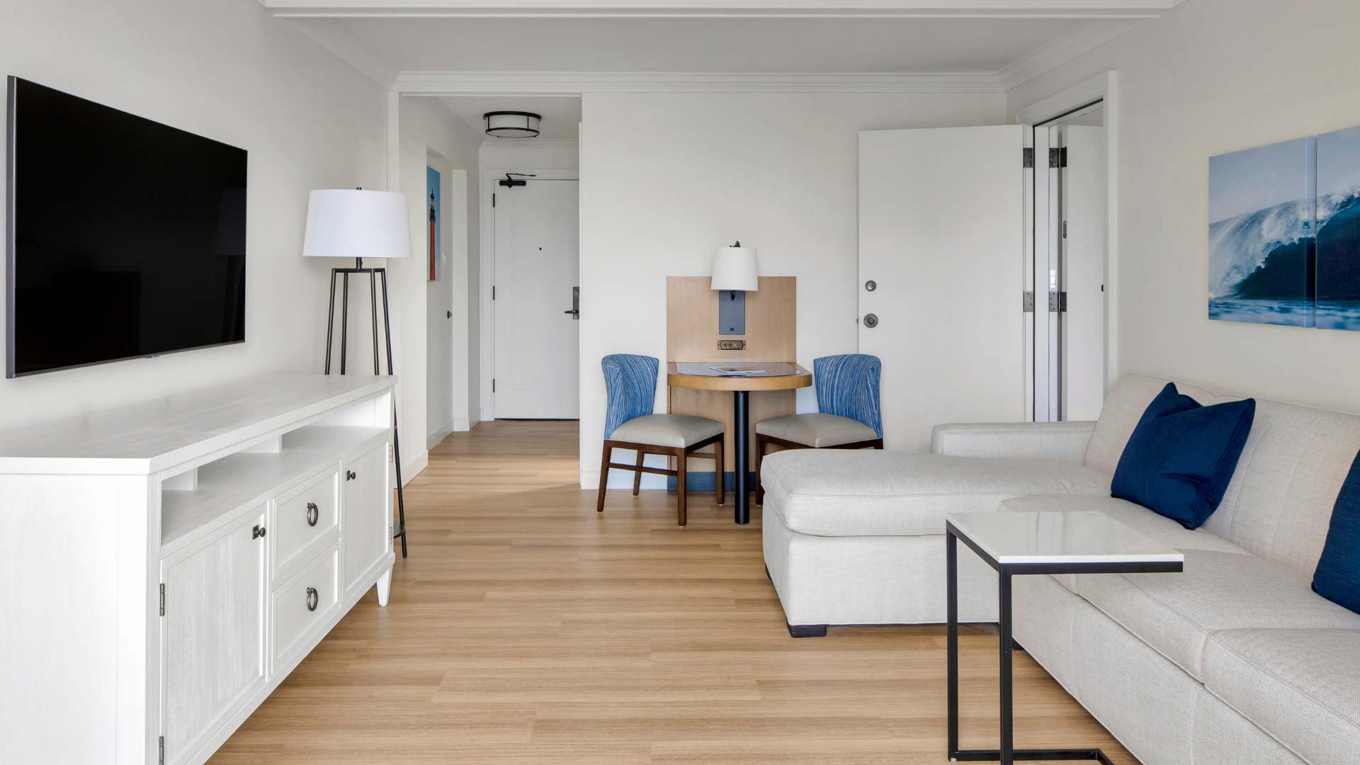 A modern living room with light wood flooring and neutral walls. It features a white entertainment console with a flat-screen TV, a beige sofa with blue cushions, a small round table with two blue chairs, and a tall lamp. A door is slightly open.