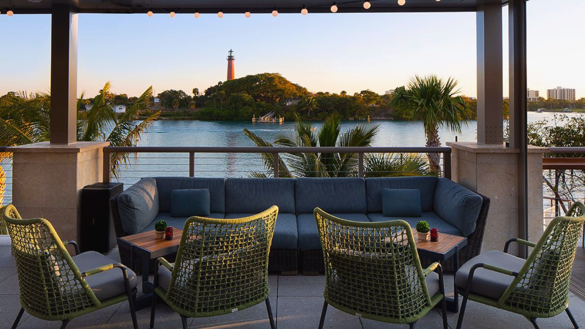Outdoor patio with wicker chairs and sofas under string lights, overlooking a calm body of water and a small, lush island. A lighthouse is visible on the island. Palm trees and city buildings appear in the distant background at sunset.