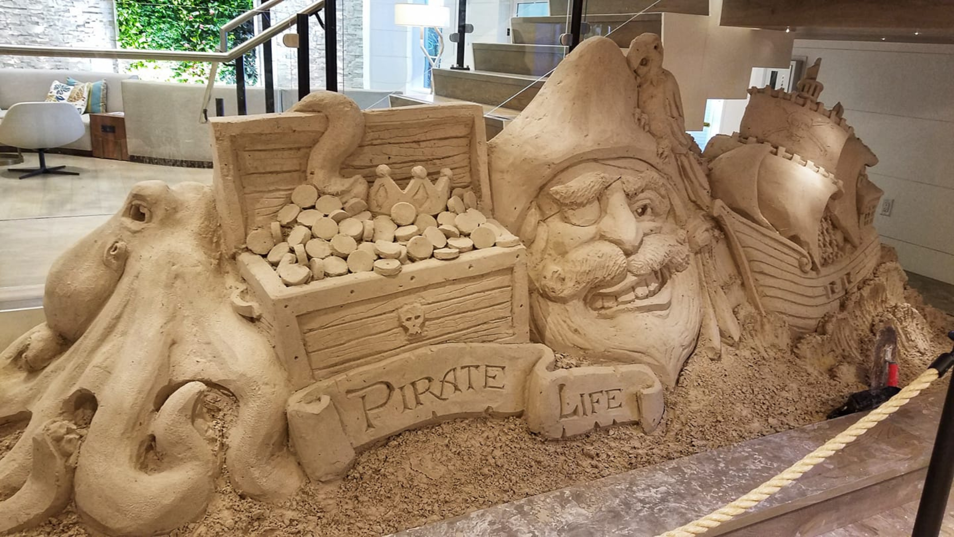 Sand sculpture featuring a pirates head, treasure chest with coins, an octopus, and a ship. The inscription Pirate Life is carved into the sand, and the scene is set indoors with stairs and greenery in the background.