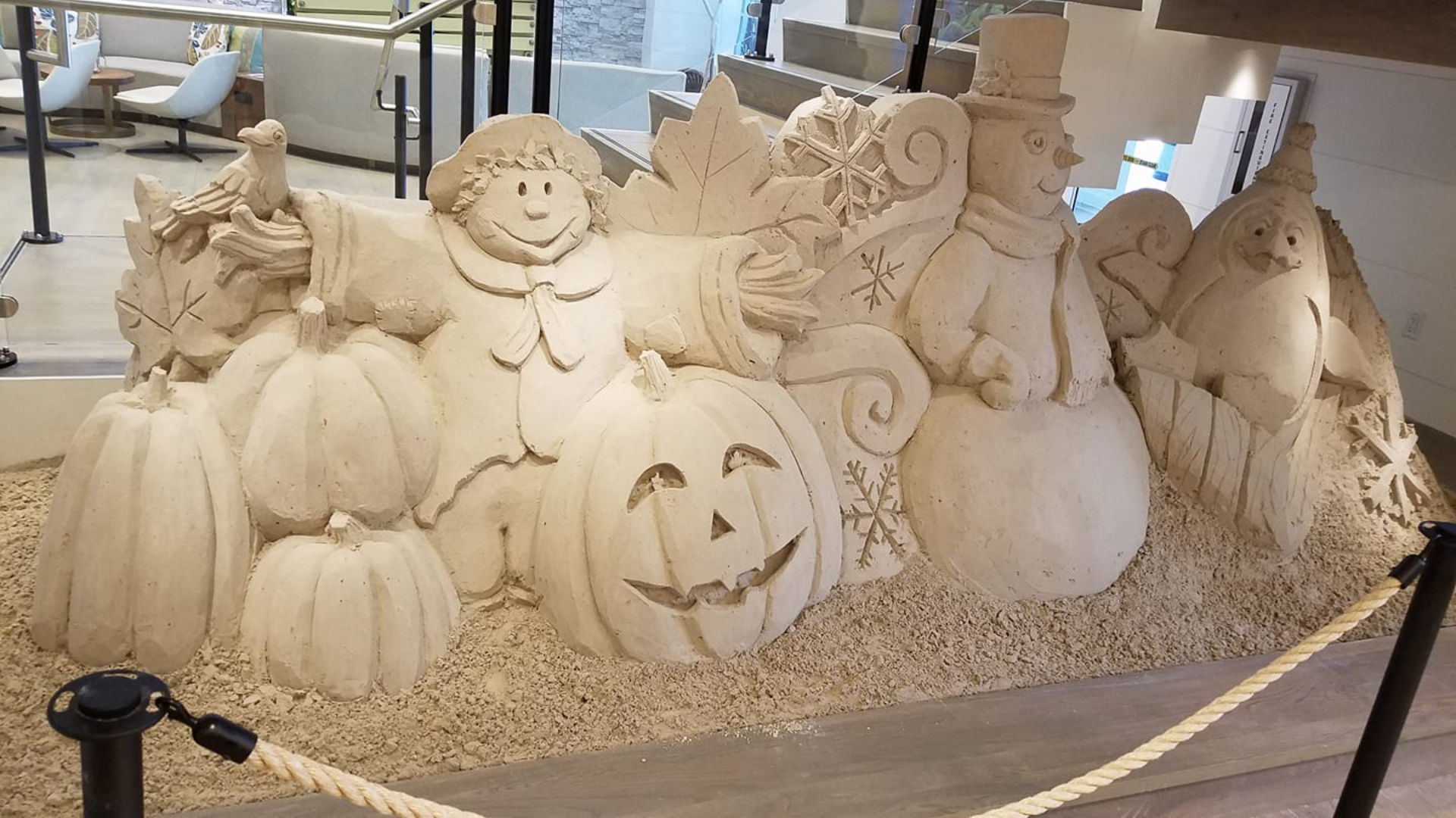 A detailed sand sculpture featuring Halloween and winter holiday themes. It includes carved pumpkins, a jack-o-lantern, cheerful scarecrows, a snowman, and snowflakes. The sculpture is surrounded by a rope barrier inside a building.