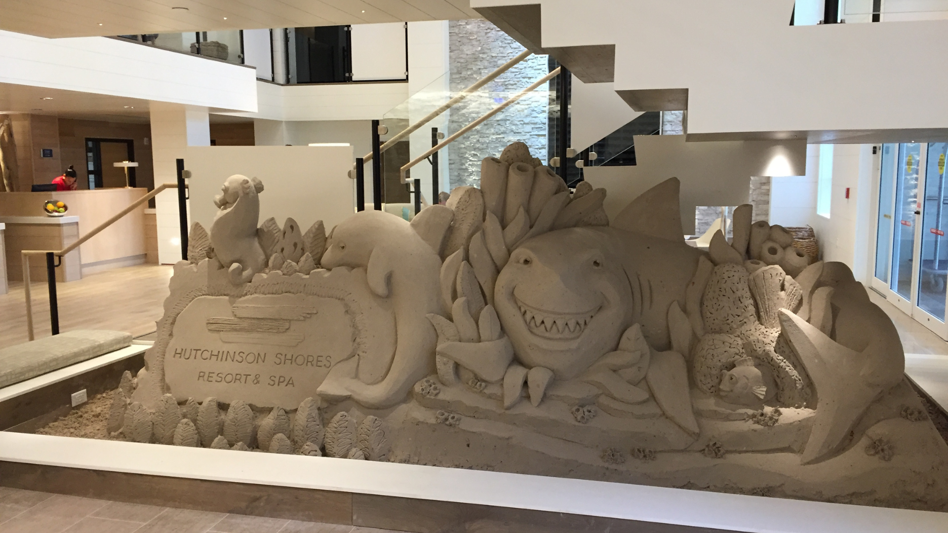 A large sand sculpture in a hotel lobby depicts sea creatures like sharks, dolphins, and fish. It features a sign that reads Hutchinson Shores Resort & Spa. The artwork is under a staircase with modern decor in the background.