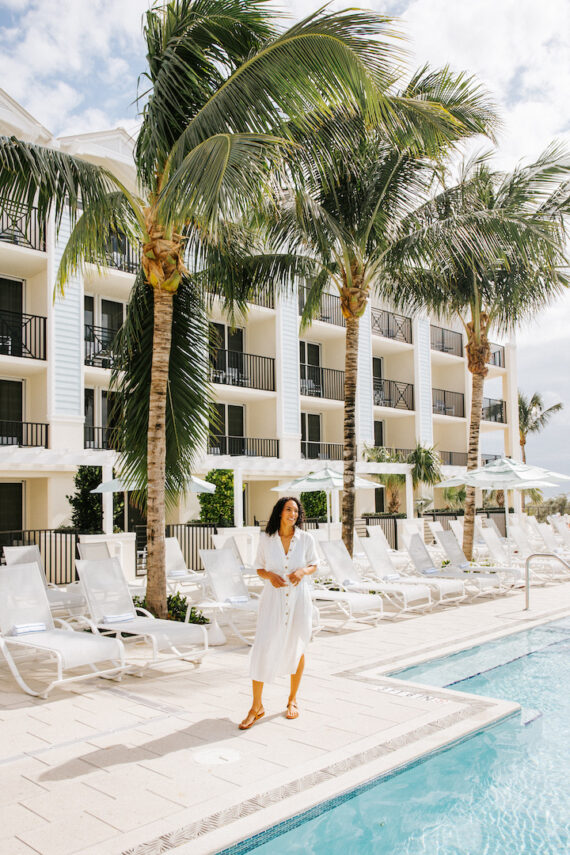 A woman in a white dress and sandals walks along the edge of a pool, surrounded by palm trees and a modern hotel with balconies. White lounge chairs line the poolside under a partly cloudy sky.