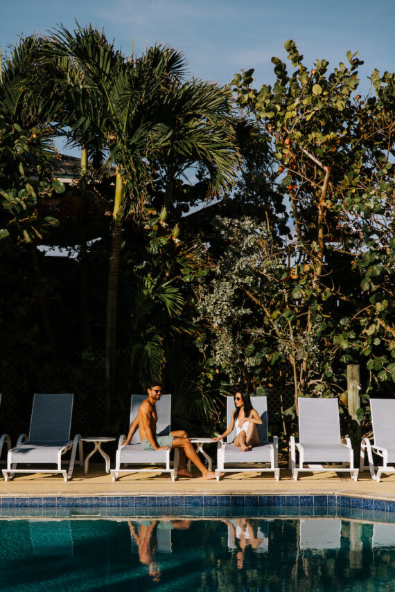 Two people sit on sun loungers by a pool, surrounded by tall palm trees and lush greenery. They are enjoying the sunny weather, and the clear blue water of the pool reflects the sky and surroundings.