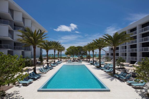 A rectangular outdoor pool lined with lounge chairs and palm trees, sits between two modern white buildings, with a view of the beach and ocean under a bright blue sky.