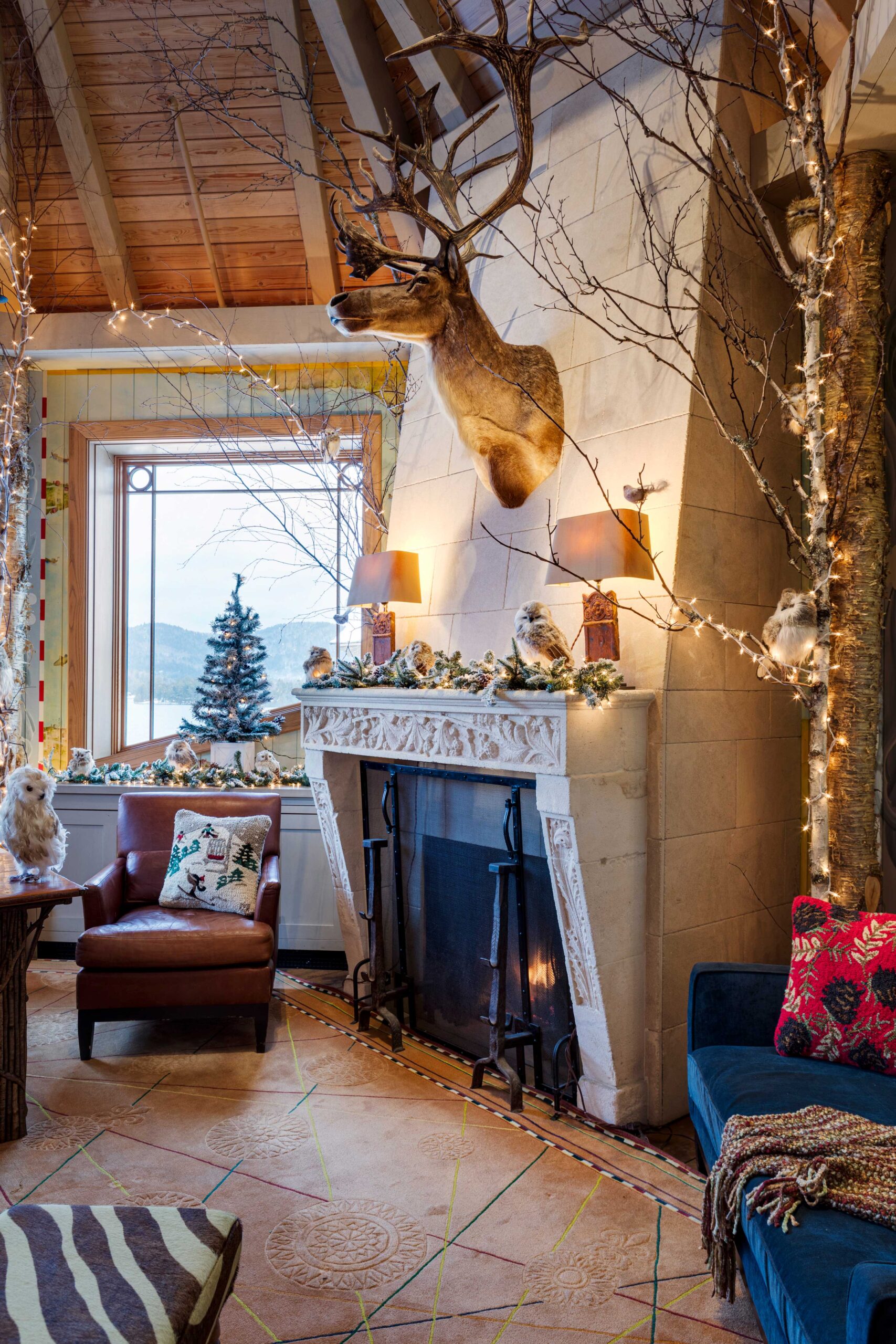Cozy living room with a stone fireplace decorated with garlands and lights, a mounted deer head above, comfy chairs, a window with a snowy view, and festive holiday decor including a small Christmas tree.