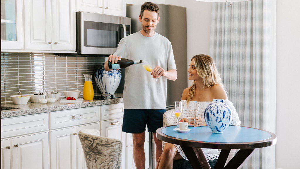 A man pours orange juice into a glass held by a smiling woman sitting at a kitchen table in a suite at Treasure Island Beach Resort. The kitchen has white cabinets, blue-and-white vases, and various breakfast items on the counter.