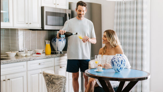 A man pours orange juice into a glass held by a smiling woman sitting at a kitchen table in a suite at Treasure Island Beach Resort. The kitchen has white cabinets, blue-and-white vases, and various breakfast items on the counter.