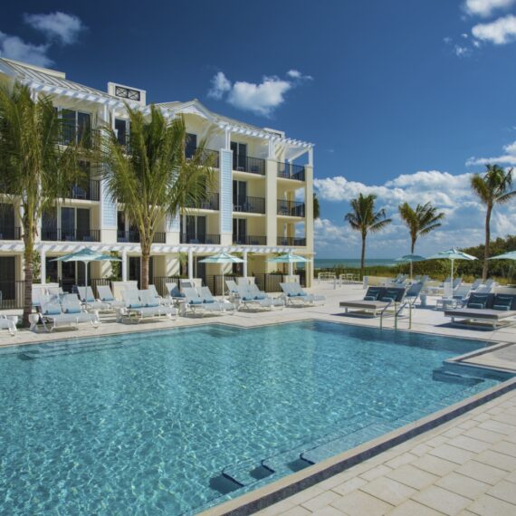 A luxurious outdoor hotel pool with blue water, surrounded by white sun loungers and palm trees. The hotel is a multi-story building with balconies. The sky is clear and sunny, suggesting a warm, inviting atmosphere.