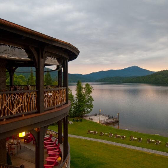 A rustic lakeside lodge with a tiered, wooden balcony featuring nature-themed railings. Adirondack chairs are arranged on the lawn, overlooking a serene lake with a distant mountain backdrop under a cloudy sky at dusk.