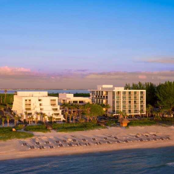Aerial view of a beachfront resort at sunset. The scene shows modern white buildings surrounded by palm trees, with sun loungers neatly arranged on the sandy beach. Gentle waves roll onto the shore, and the sky is a gradient of pink and blue.