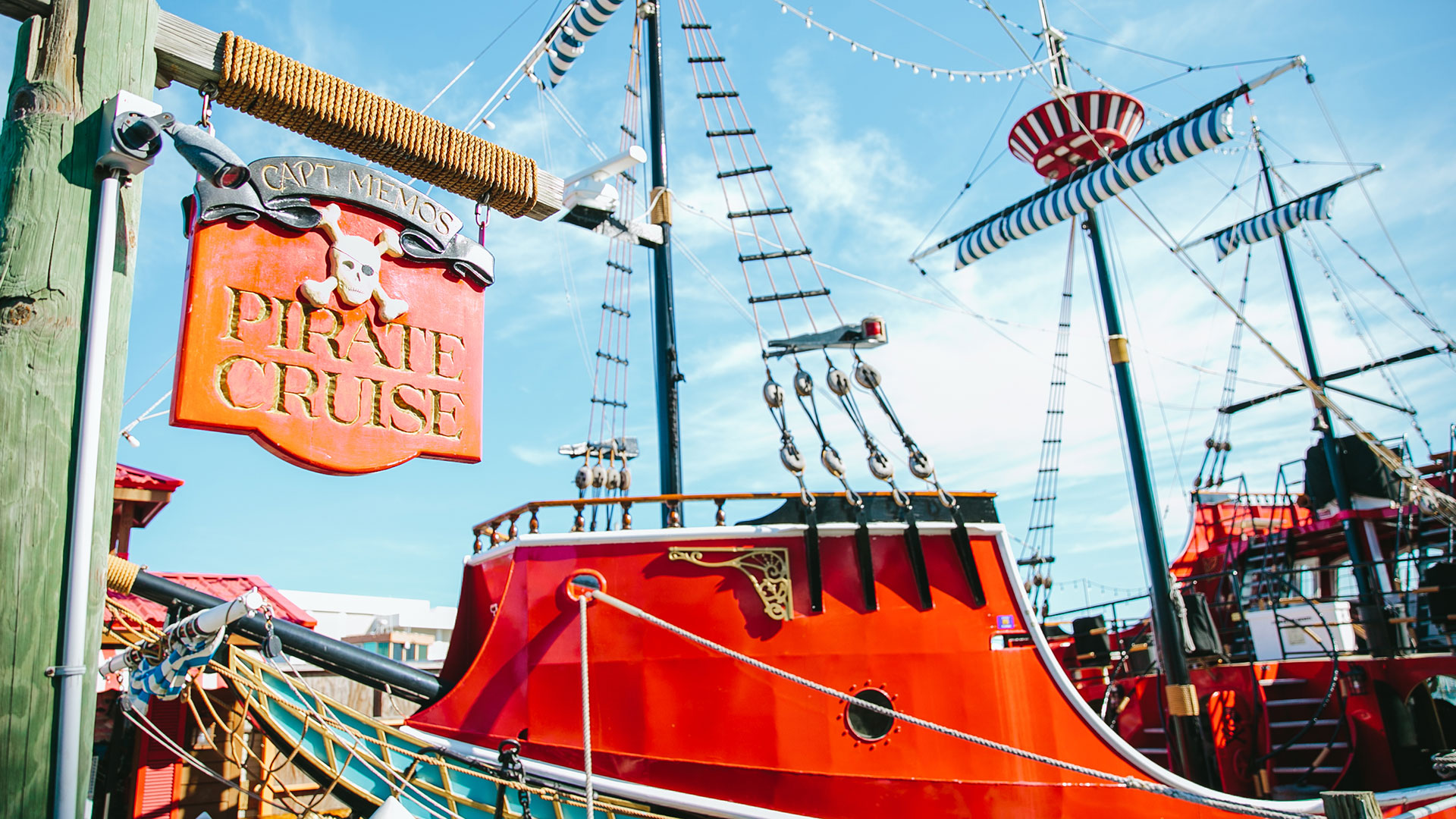 A vibrant red pirate ship with striped sails docked at a port. A wooden sign in the foreground reads Pirate Cruise with a skull emblem. The sky is clear and blue.