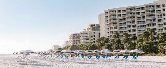 A beach lined with blue and white striped umbrellas and matching lounge chairs. In the background, tall white resort buildings rise above palm trees under a clear sky.