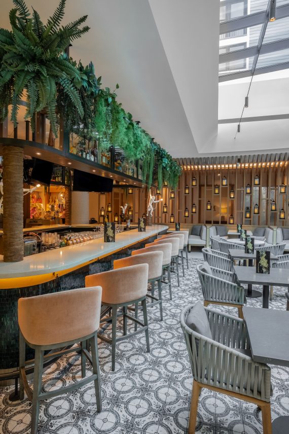 A modern restaurant interior with a wooden bar, lined with beige stools under hanging greenery. Tables with woven chairs are on a patterned tile floor. Warm lighting fills the space from candles and overhead lights. A skylight illuminates the area.