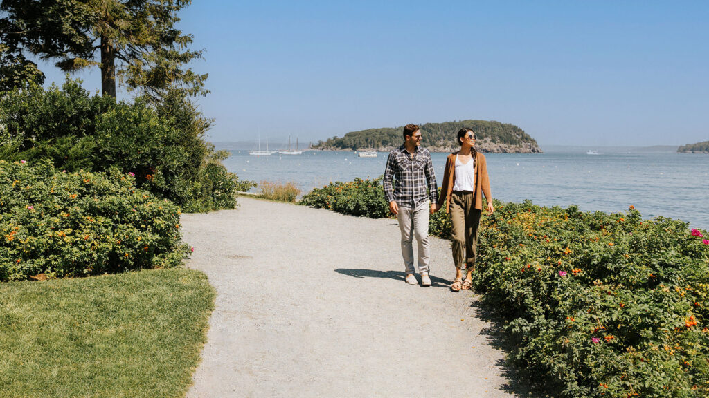 A couple walks along a gravel path by the water, surrounded by green bushes and trees; sailboats and a small island are visible in the distance under a clear blue sky, perfect for discovering fun facts about Maine’s scenic beauty.