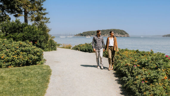 A couple walks along a gravel path by the water, surrounded by green bushes and trees; sailboats and a small island are visible in the distance under a clear blue sky, perfect for discovering fun facts about Maine’s scenic beauty.