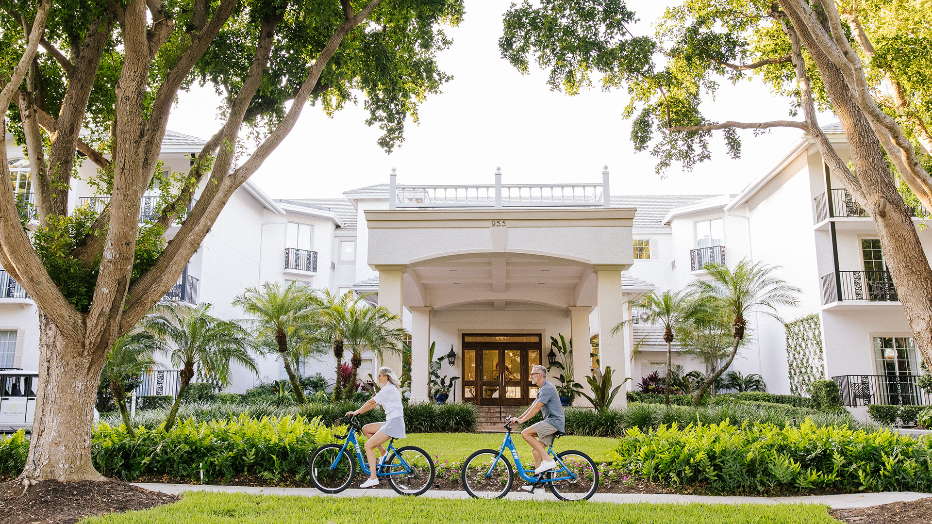 Two people ride blue bicycles on a path in front of a large, white, three-story building surrounded by lush greenery and tall trees on a sunny day.
