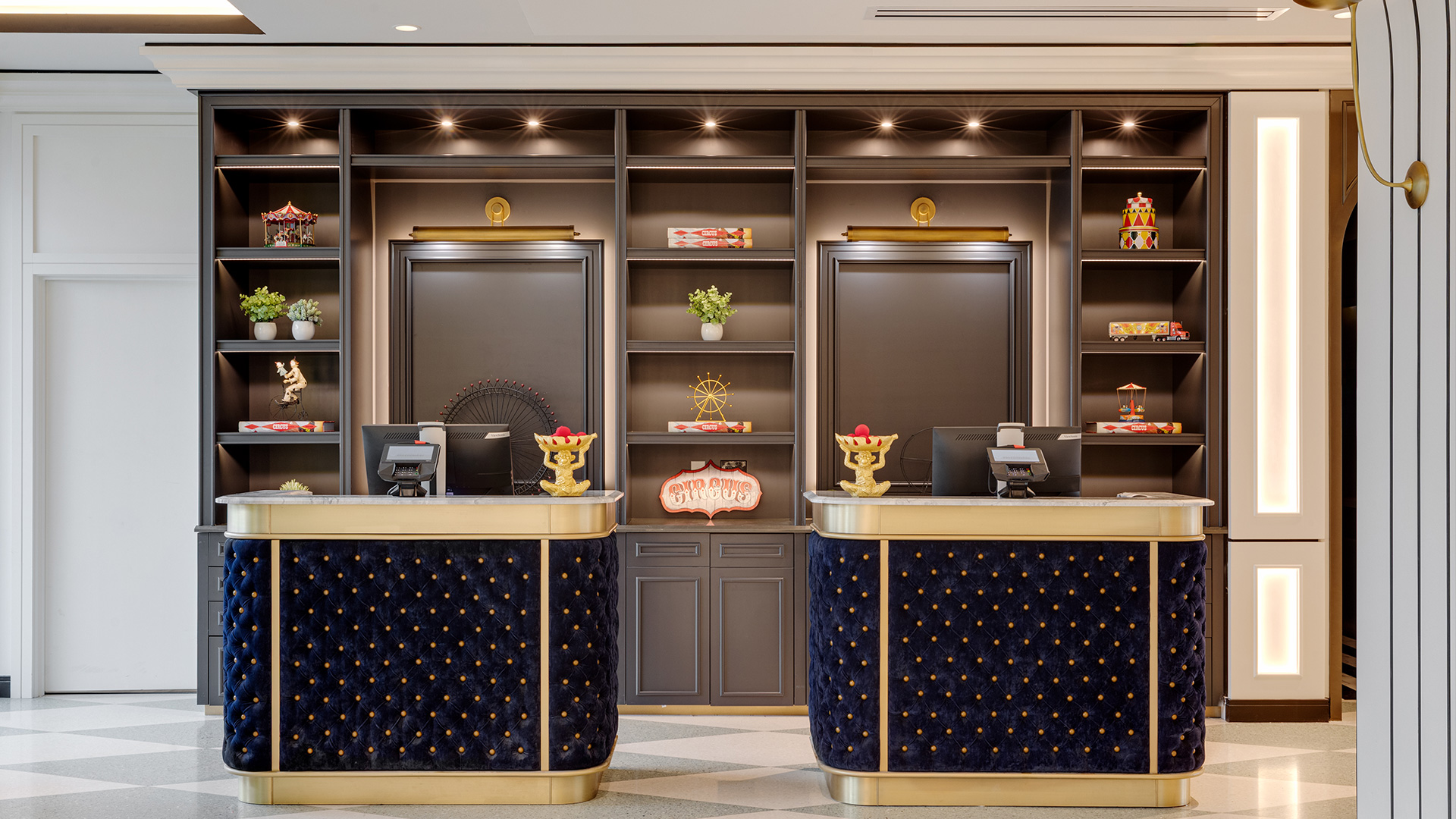 Hotel reception area with two blue and gold front desks, computer monitors, and decorative shelving behind. Shelves display plants, figurines, and small objects—warm lighting highlights the elegant decor found at Hotels On Lido Beach Sarasota.