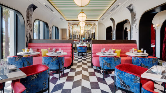 Elegant restaurant interior near Hotels On Lido Beach Sarasota features pink booth seating, blue patterned chairs, and a black-and-white checkered floor. Arched windows and modern hanging lights enhance the bright, stylish dining atmosphere.