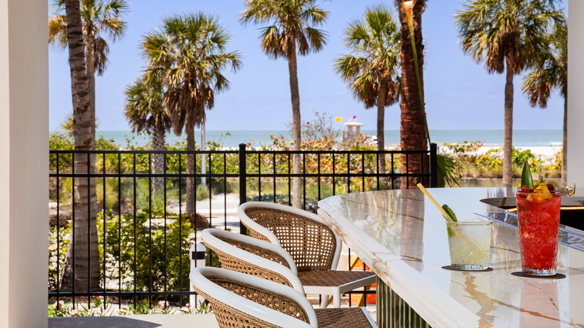 A marble outdoor bar with wicker chairs overlooks a beach lined with palm trees. Two colorful cocktails sit on the bar, capturing the relaxed vibe of Hotels On Lido Beach Sarasota, with ocean views and a bright blue sky in the background.