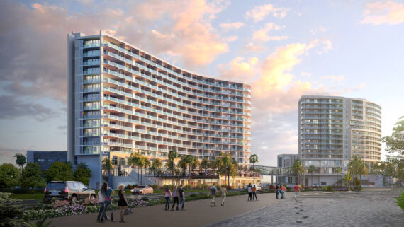 A modern, curved high-rise building with glass balconies surrounded by palm trees under a partly cloudy sky. People are walking on the plaza, and a car is parked nearby. Another circular building is visible in the background.