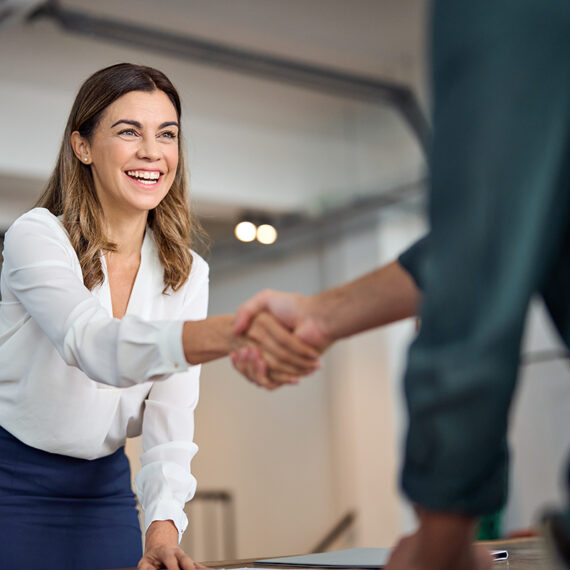 A woman in a white blouse is smiling and shaking hands across a table with a person wearing a green shirt. The background is an office setting with blurred lights and furniture.
