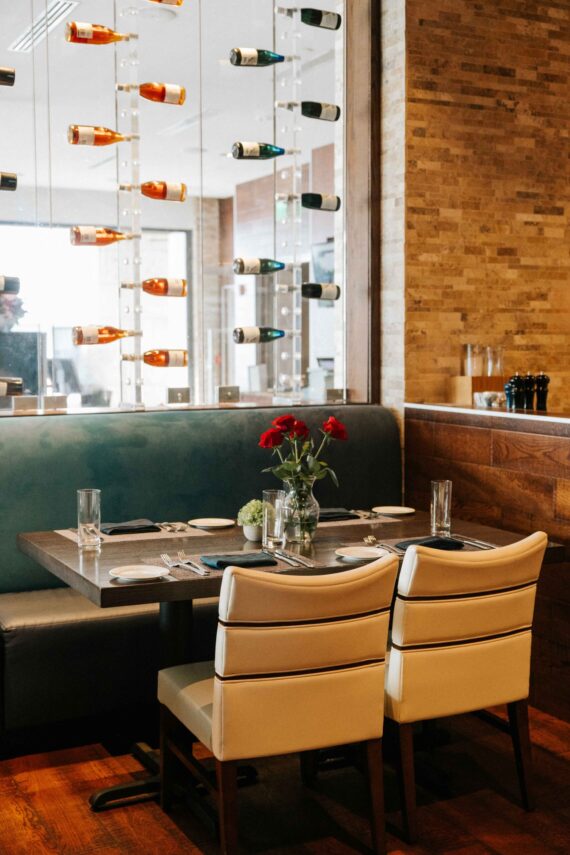 A cozy restaurant booth with a wooden table set for two, featuring white chairs. The table has a vase with red roses and a small plant. A glass wall displays various wine bottles. The decor includes warm-toned wood accents.