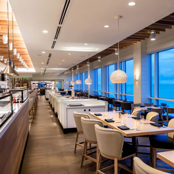 Modern restaurant interior with wooden flooring, white tables, and beige chairs. Pendant lights hang from the ceiling. Large windows on the right offer a view of the sky. The space has a clean and elegant design.