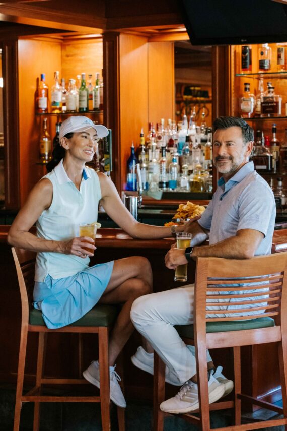 A woman and a man are sitting at a bar with wooden decor, holding glasses of beer. The woman is wearing a visor, sleeveless shirt, and skirt, while the man is in a light shirt and pants. The bar is stocked with various bottles.