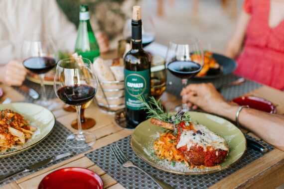 A wooden table is set with plates of pasta dishes, red wine glasses, and a bottle of wine. A main dish featuring meat topped with cheese and herbs is in focus. People are seated around the table, enjoying the meal.
