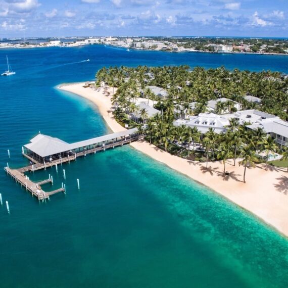 Aerial view of a coastal area with turquoise waters and a sandy beach lined with palm trees. Several buildings are nestled among the trees. A long pier extends into the water, and a small sailboat is visible in the distance.