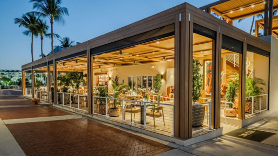 Outdoor view of a modern, well-lit restaurant with open sides, wooden beams, lush plants, and stylish tables and chairs, set on a brick-paved sidewalk under palm trees at dusk.