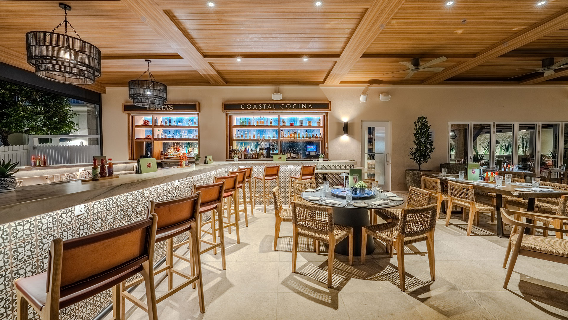 Modern restaurant interior with a long bar lined with wooden stools, tables set for dining, patterned tile accents, warm lighting, and shelves stocked with bottles behind the bar labeled “Coastal Cocina.”.