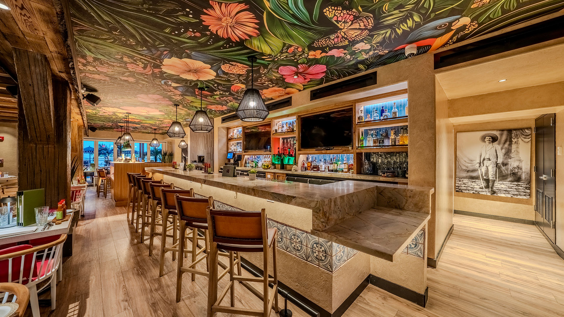 A stylish restaurant bar features wooden chairs lined along a tiled counter, shelves with colorful bottles, and a vibrant floral ceiling mural. Warm lighting and rustic decor create a cozy, inviting atmosphere.