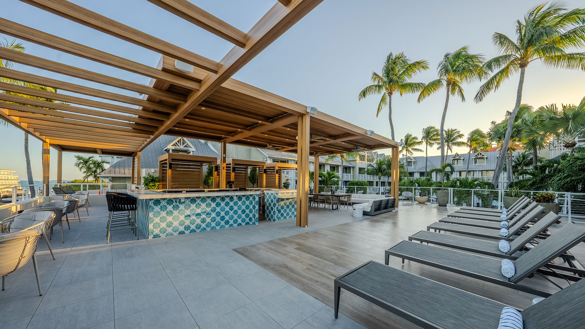 Outdoor lounge area with a wooden pergola, bar with blue tile accents, rows of lounge chairs, and palm trees, overlooking a resort with buildings in the background under a clear sky.