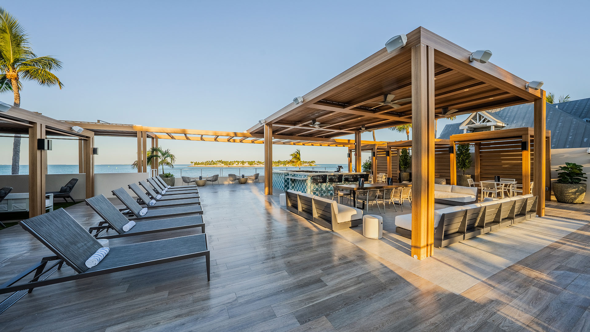 Modern outdoor lounge area with wooden cabanas, sun loungers, and a bar overlooking the ocean. The area is spacious, with palm trees and clear skies, creating a relaxing, tropical atmosphere.