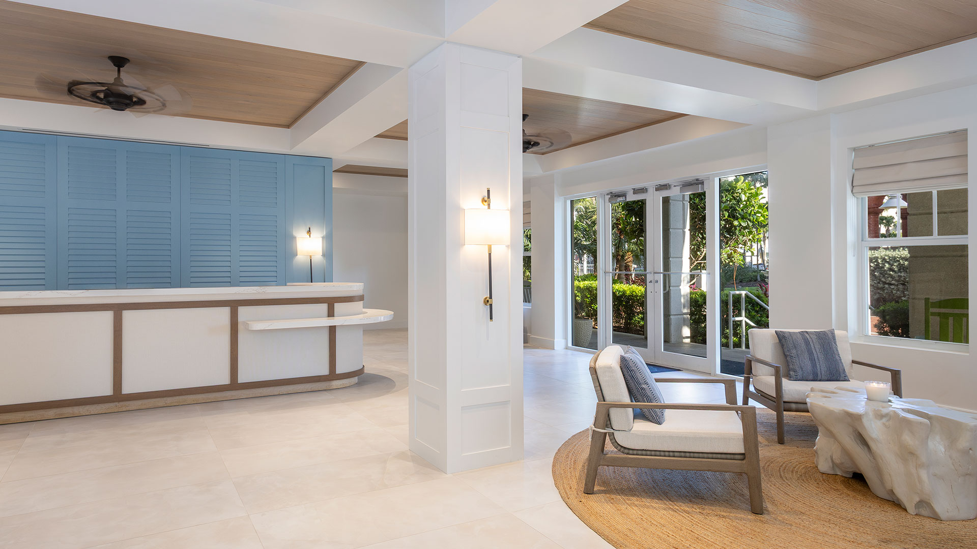 Opal Key's new modern, airy lobby with light tile floors, a curved reception desk with blue paneling, two cushioned chairs on a woven rug, and large windows letting in natural light with views of greenery outside.