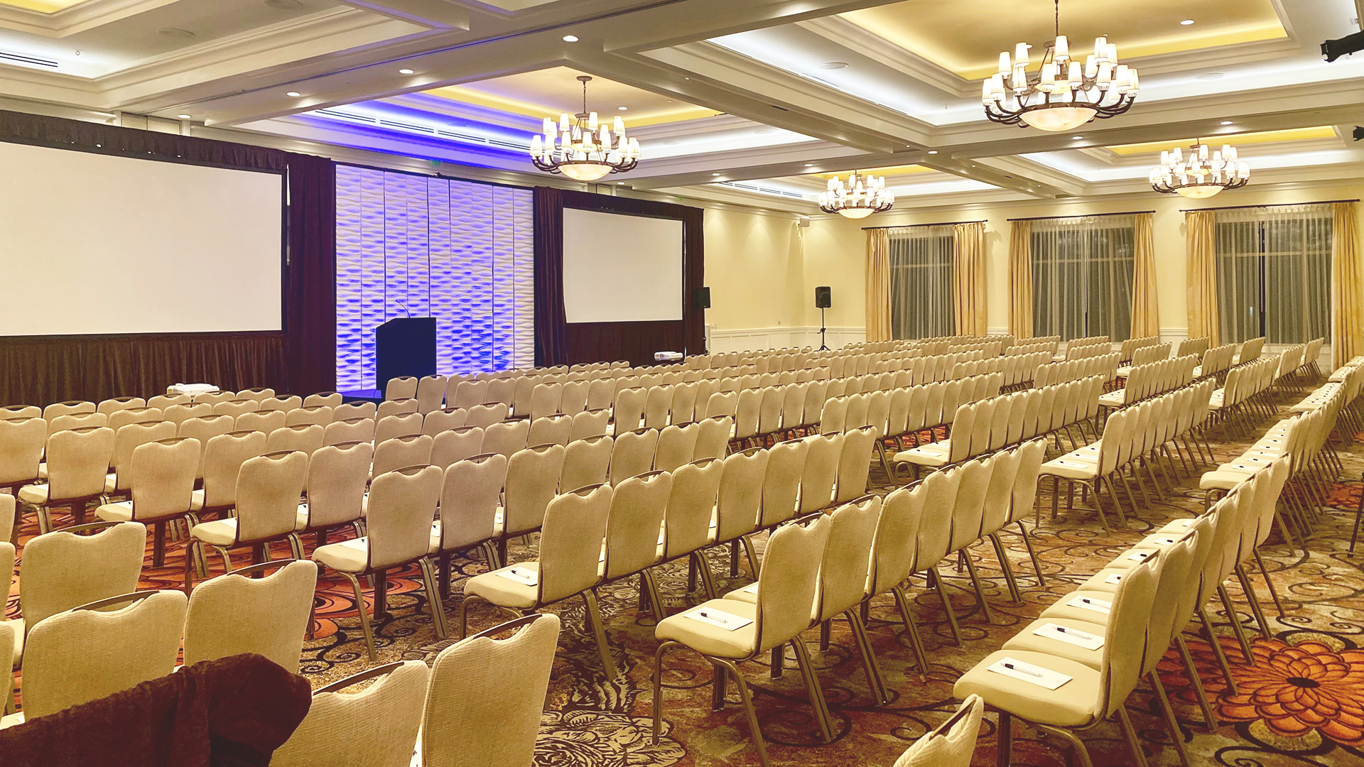An empty conference room with rows of beige chairs facing two large projection screens. The room has a patterned carpet, chandeliers, and ambient lighting. A podium is placed near the screens.
