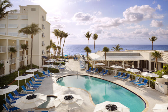 The tropical beachfront view at Delray Sands Resort in Delray, Florida, that features a curvy pool surrounded by blue lounge chairs and white umbrellas. Tall palm trees line the area, and the ocean is visible in the background under a partly cloudy sky.