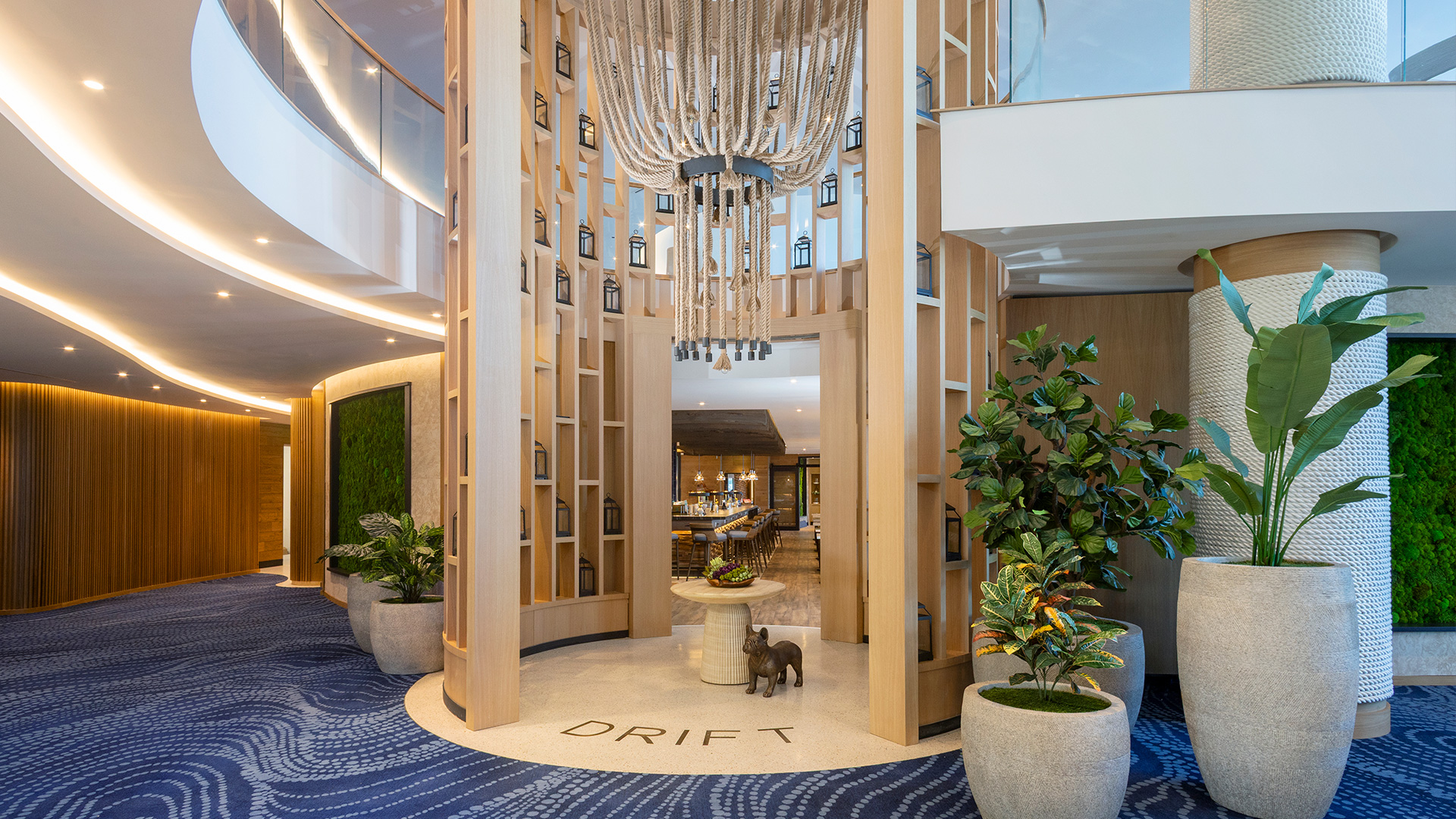 Modern hotel lobby with blue patterned carpet, tall wooden panels, large potted plants, a chandelier, and a round table with decorative items in the center. The word DRIFT is written on the floor.