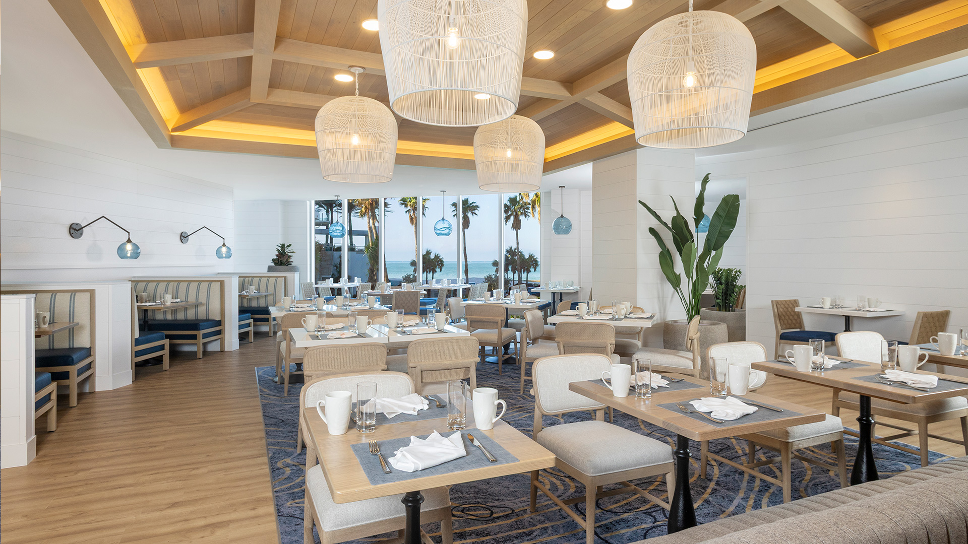 A bright, modern restaurant with wooden tables, cushioned chairs, and booth seating. Large wicker pendant lights hang from a wooden ceiling. Palm trees and the ocean are visible through large windows at the back.