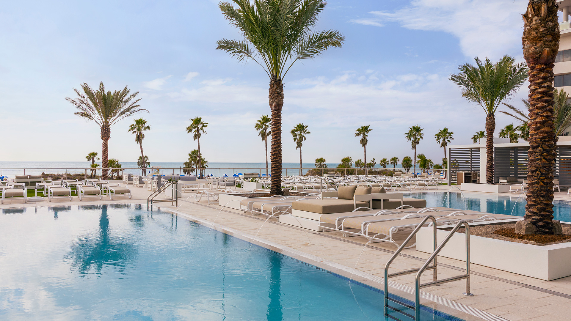 A modern outdoor swimming pool with lounge chairs and palm trees overlooks a sandy beach and the ocean on a sunny day.