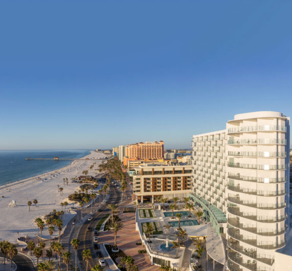 Aerial view of a modern beachfront hotel and palm-lined road beside a wide sandy beach, with clear blue sky and calm ocean stretching into the distance.