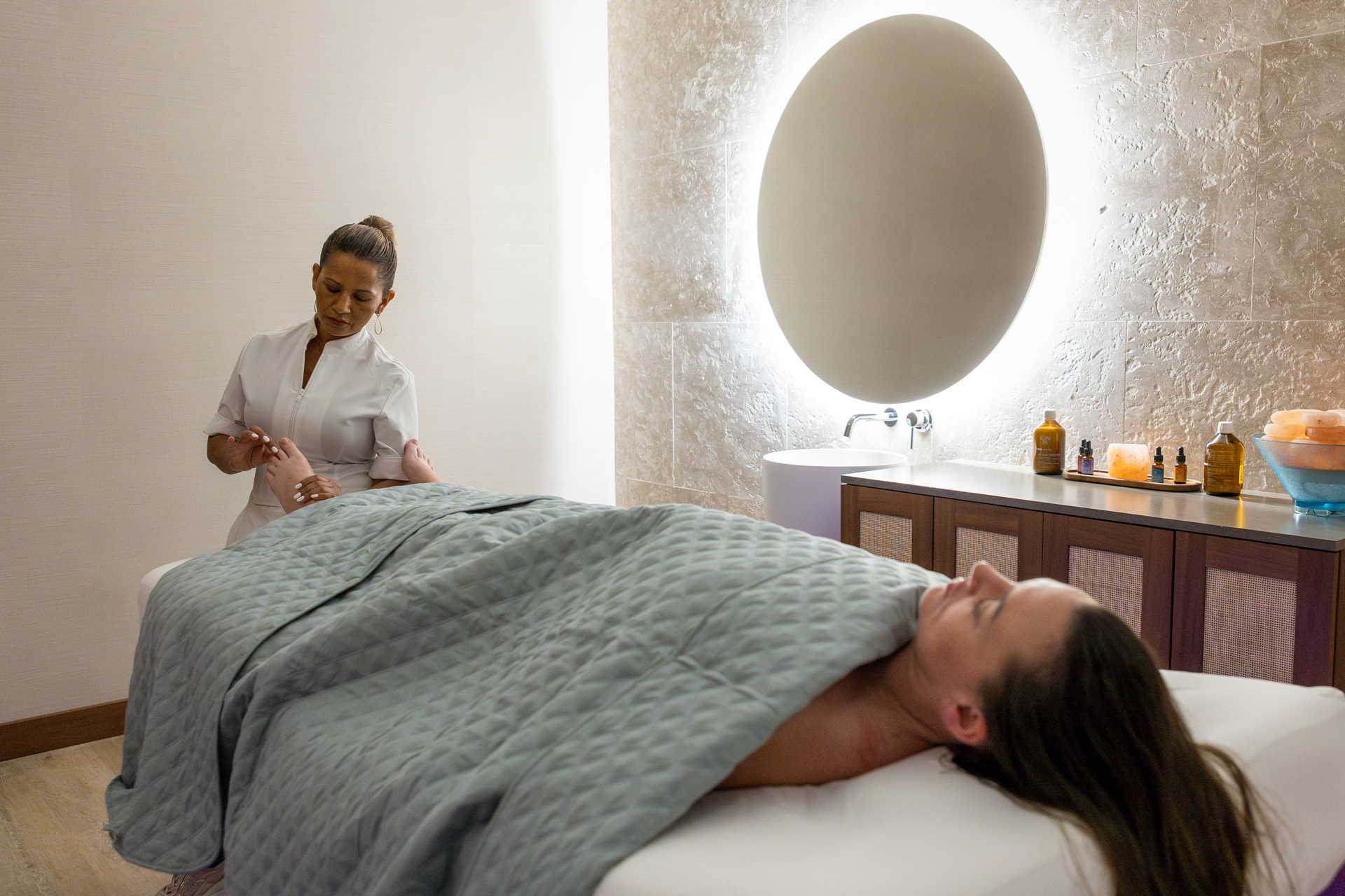 A therapist in white stands at the head of a woman on a treatment table, mirror and stone wall behind.