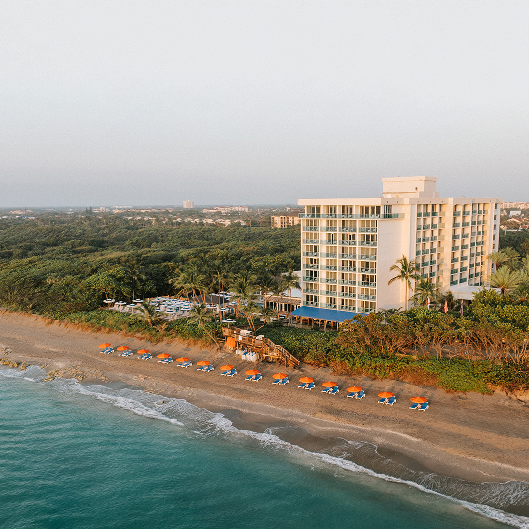 A beachfront hotel with multiple floors sits beside the ocean, surrounded by lush green trees. A row of orange and blue beach umbrellas lines the sandy shore near the turquoise water.