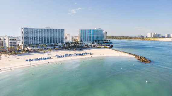 Aerial view of a beach with rows of blue lounge chairs and new hotels in Clearwater Beach, Florida, bordering the ocean.