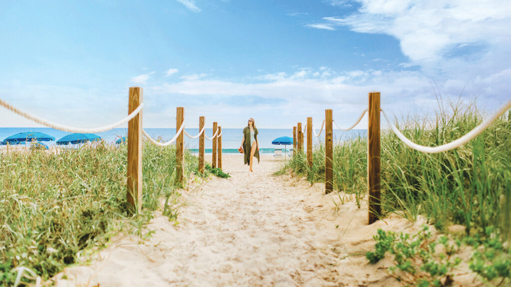 A woman in a green dress walks along a sandy path lined with rope and posts, leading to the best beach Florida has to offer, with blue umbrellas, green grass on either side, and a bright blue sky dotted with clouds.