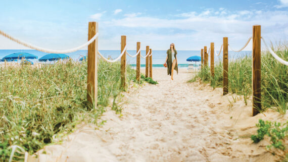 A woman in a long dress walks on a sandy path lined with rope and wooden posts toward the best beach Florida has to offer, where blue umbrellas and lounge chairs await under a bright, partly cloudy sky.